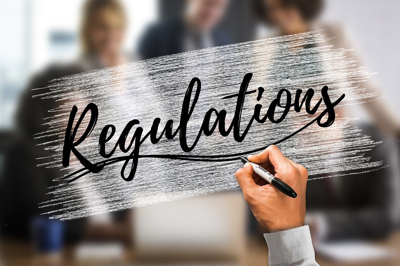 office-4249390_1280 A hand holding a marker writes the word 'Regulations' in elegant script on a transparent surface, with a blurred background of business professionals in a meeting.